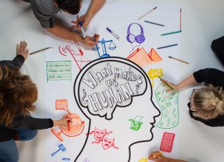 What Makes Us Human? People are seen from above drawing objects on a poster with markers and colored pencils
