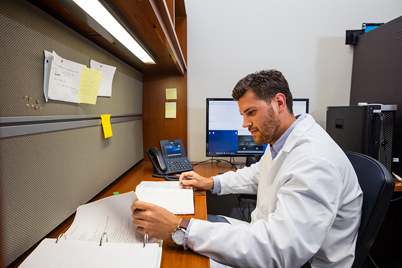 A student wearing a white lab coat sits at a desk and flips through a 3-ring binder A student wearing a white lab coat sits at a desk and flips through a 3-ring binder